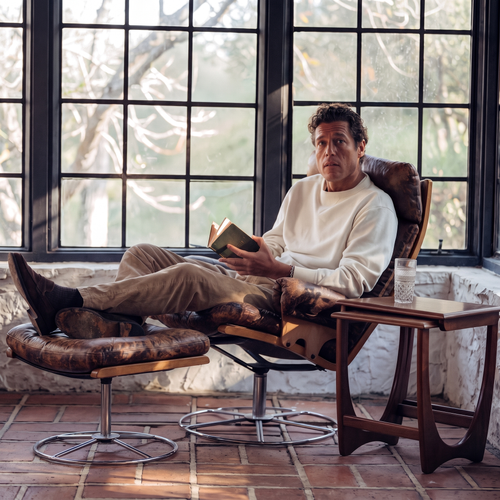 A man in a cream sweater reads a book in a leather and bentwood swivel chair with his feet up on a matching footstool, beside a tall black-framed window