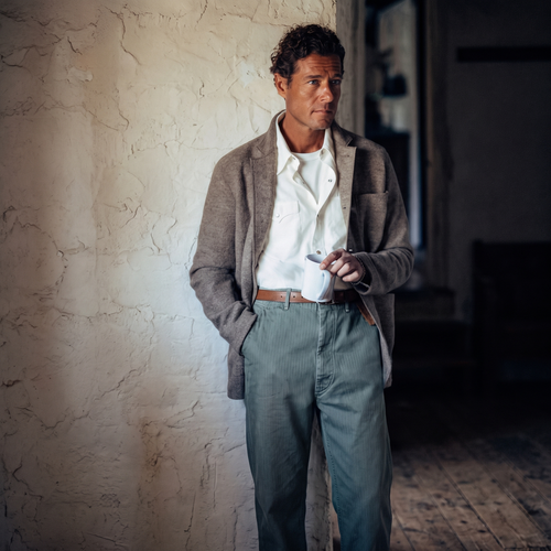 A man in a grey wool coat over a white shirt stands against a cream stucco wall, holding a mug in one hand.
