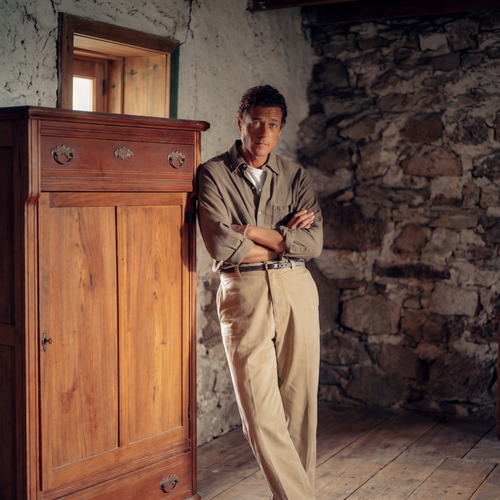 A man in a khaki overshirt and light trousers stands with his arms crossed beside a tall wooden cabinet, in front of a rough stone wall.
