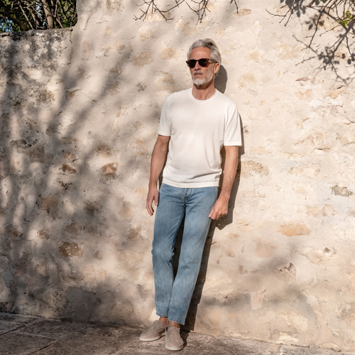 A grey-haired man in a white T-shirt, blue jeans and sunglasses stands against a pale stone wall in dappled sunlight.
