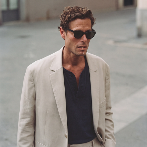 A man in a cream blazer over a dark navy shirt, wearing sunglasses with a matchstick in his mouth, on a pale sunlit street.
