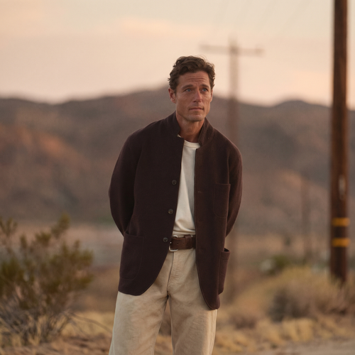 A man in a dark brown wool chore jacket over a cream shirt and cream trousers stands outdoors at dusk, with blurred hills behind him.