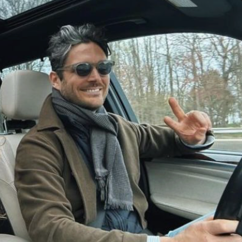 A man in a green suede jacket and grey scarf, wearing sunglasses, sits smiling in the driver's seat of a car with bare trees visible through the window.