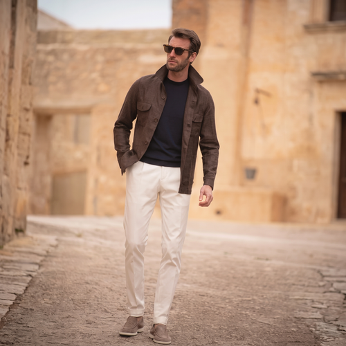 A man in a brown overshirt, black T-shirt and white trousers walks across sand-coloured ground beside old stone buildings.